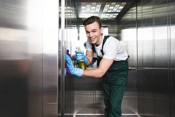 Hübsche junge Reinigungskraft putzt Fahrstuhl und lächelt in die Kamera — Stockfoto