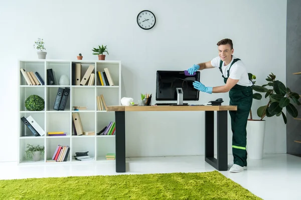 Hübsche junge Putzfrau lächelt in die Kamera, während sie im Büro Computer putzt — Stockfoto