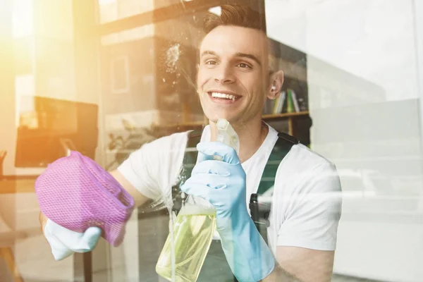 Guapo joven sonriente limpiando y limpiando la ventana con aerosol botella y trapo - foto de stock