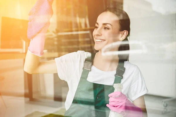 Hermosa mujer joven sonriente limpiando y limpiando la ventana con aerosol botella y trapo - foto de stock