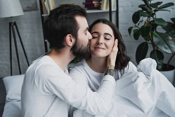 Bello uomo barbuto baciare bella donna sorridente a casa a letto — Foto stock