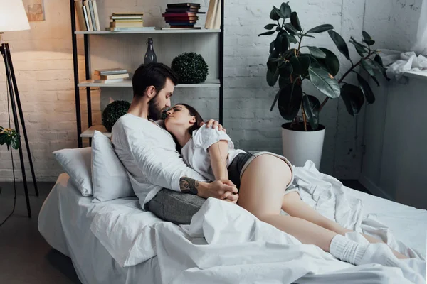 Beau jeune couple tenant la main et embrassant tout allongé dans le lit à la maison — Photo de stock