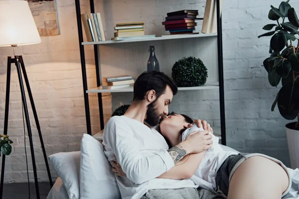 Hermosa pareja joven abrazos y besos mientras está acostado en el dormitorio - foto de stock