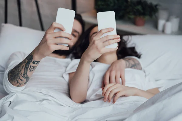 Jeune couple couché au lit et utilisant des smartphones — Photo de stock