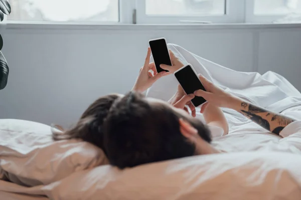 Pareja joven acostada en la cama y utilizando teléfonos inteligentes con pantalla en blanco - foto de stock