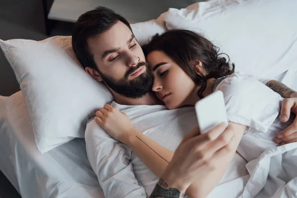 Homem bonito usando smartphone enquanto mulher dormindo na cama — Fotografia de Stock