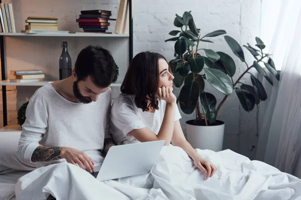 Bärtiger Mann liegt im Bett und benutzt Laptop, während gelangweilte Frau ihr Kinn im Schlafzimmer stützt — Stockfoto