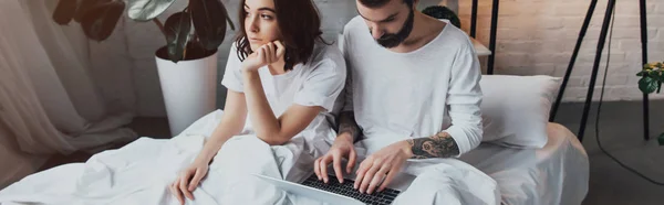 Barbudo hombre acostado en la cama y el uso de la computadora portátil, mientras que la mujer aburrida apoyo barbilla en el dormitorio - foto de stock