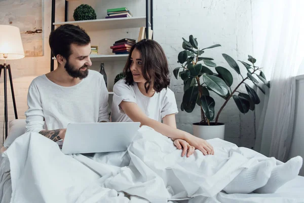 Hermosa pareja joven en pijama acostado en la cama, utilizando el ordenador portátil y mirándose - foto de stock