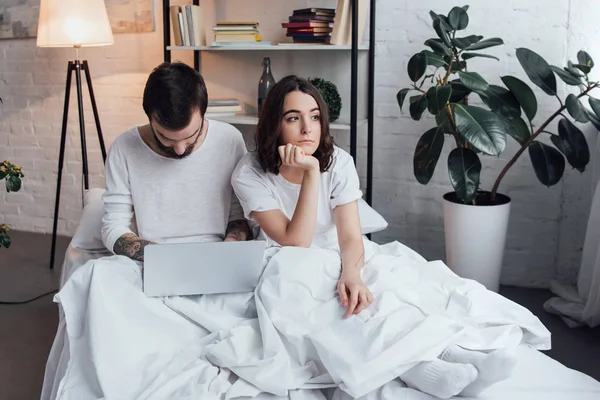 Hombre acostado en la cama y el uso de la computadora portátil, mientras que la mujer aburrida apoyo barbilla y mirando hacia otro lado en el dormitorio - foto de stock