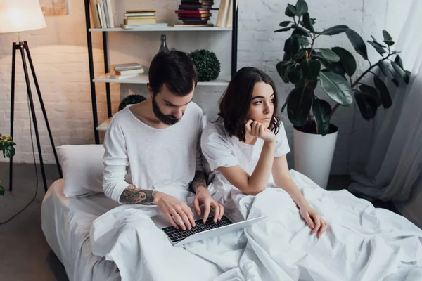 Uomo sdraiato a letto e utilizzando il computer portatile mentre la donna annoiata puntamento mento e guardando lontano in camera da letto — Foto stock