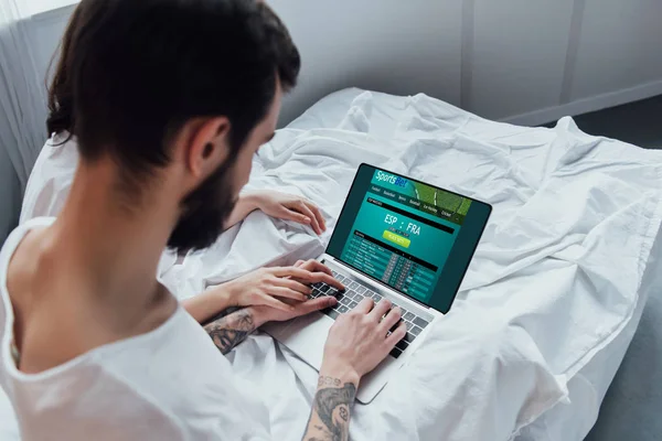 Задний вид пары, лежащей в постели, держась за руки и используя ноутбук со спортивным сайтом на экране — стоковое фото