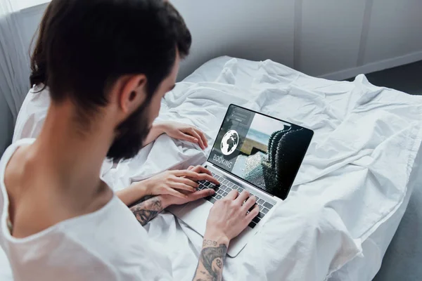 Vista trasera de la pareja acostada en la cama, tomados de la mano y utilizando el ordenador portátil con el sitio web de reserva en pantalla - foto de stock