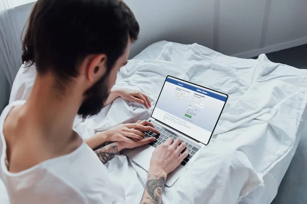 Back view of couple lying in bed, holding hands and using laptop with facebook website on screen — Stock Photo