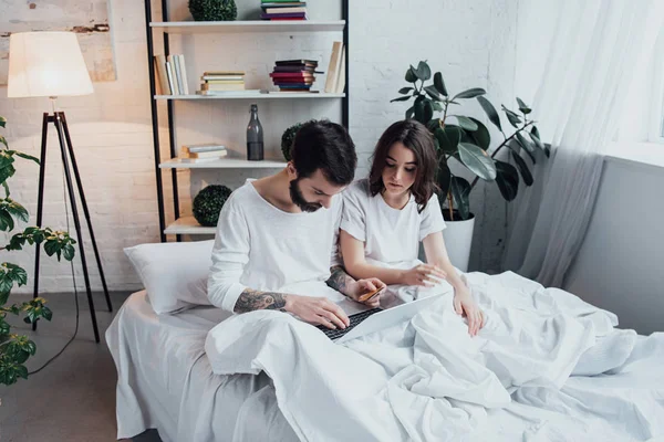 Belo jovem casal de pijama deitado na cama e usando laptop — Fotografia de Stock