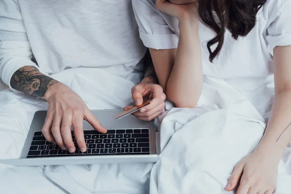Vista recortada de la pareja tatuada con tarjeta de crédito y portátil tumbado en la cama y haciendo compras en línea - foto de stock