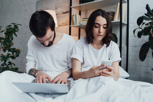 Hermosa pareja joven en pijama acostado en la cama mientras se utiliza el ordenador portátil y teléfono inteligente - foto de stock