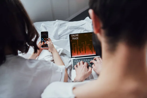 Вид ззаду пари лежить в ліжку і використовує цифрові пристрої з онлайн-торгівлею і графіками на екрані — стокове фото
