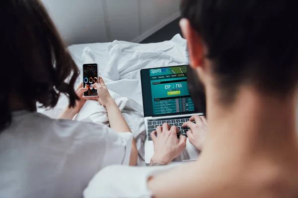 Vue arrière du couple couché au lit et utilisant des appareils numériques avec paris sportifs et graphiques à l'écran — Photo de stock