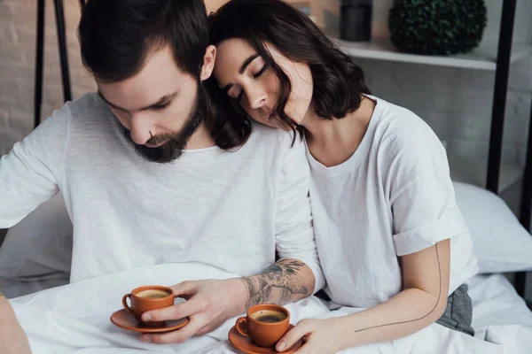 Beautiful young tattooed couple lying in bed with coffee cups in morning — Stock Photo