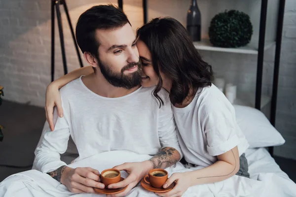 Schönes junges Paar, das morgens mit Kaffeetassen im Bett liegt und sich umarmt — Stockfoto