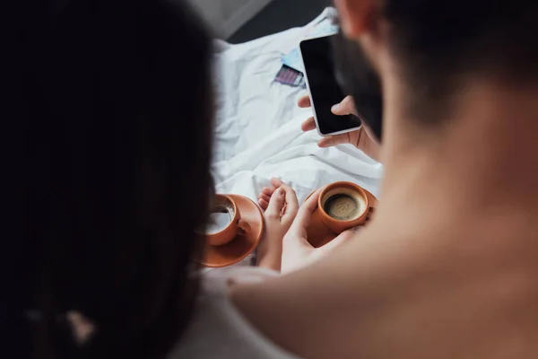 Back view of couple with coffee cups using smartphone with blank screen at home — Stock Photo