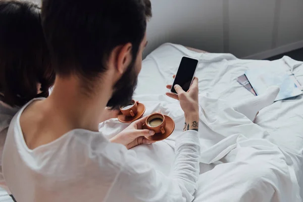 Rückansicht eines jungen Paares, das morgens mit Kaffee im Bett liegt und Smartphone mit leerem Bildschirm benutzt — Stockfoto