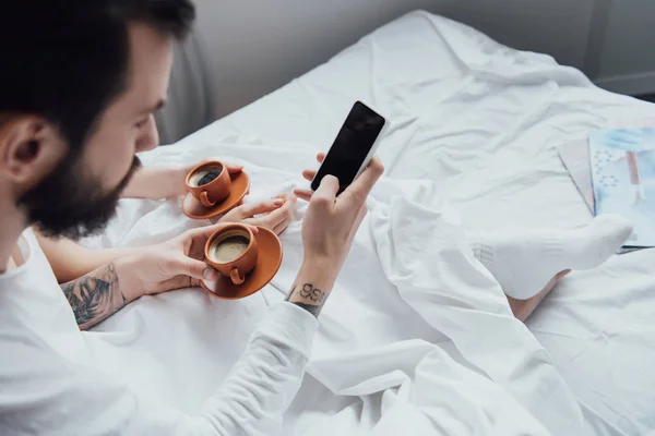 Vista recortada de la joven pareja acostada en la cama con café y el uso de teléfono inteligente con pantalla en blanco por la mañana - foto de stock