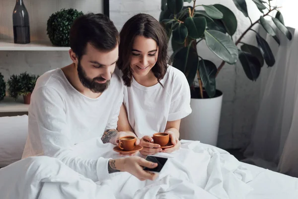 Bella giovane coppia sdraiata a letto con il caffè e utilizzando lo smartphone al mattino — Foto stock