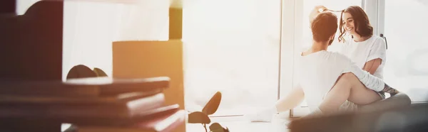 Селективный фокус красивой молодой пары, обнимающей и сидящей дома на подоконнике с копировальным пространством и солнечным светом — стоковое фото