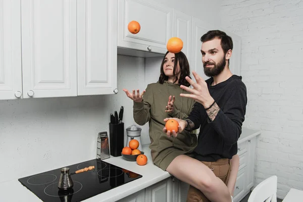 Belo jovem casal se divertindo e malabarismo com laranjas na cozinha — Fotografia de Stock