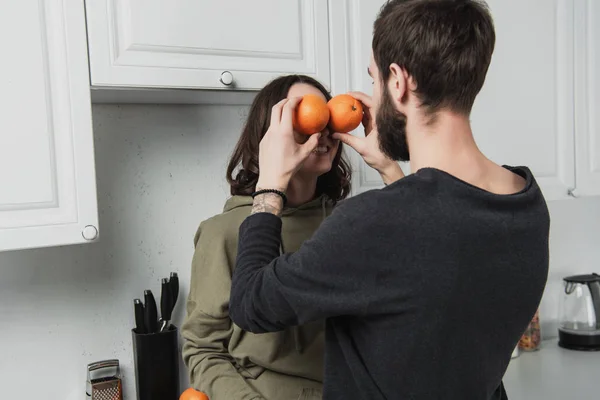 Vista trasera del hombre sosteniendo naranjas delante de la cara de la mujer en la cocina - foto de stock