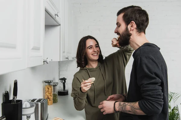 Belle femme souriante nourrissant bel homme avec des toasts pendant le petit déjeuner dans la cuisine — Photo de stock