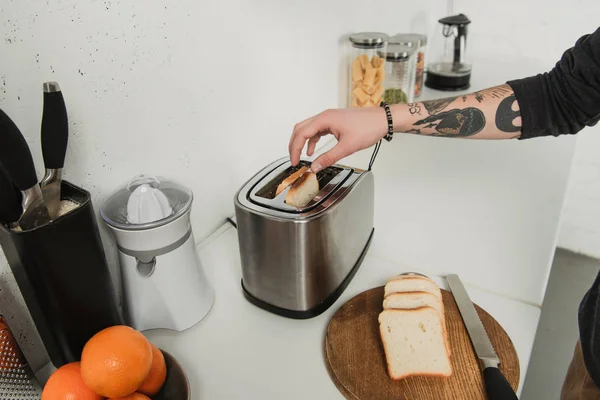 Cropped view of tattooed man preparing toasts with toaster during breakfast in kitchen — Stock Photo