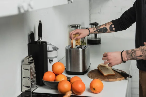 Vista recortada del hombre tatuado preparando tostadas con tostadora durante el desayuno en la cocina - foto de stock