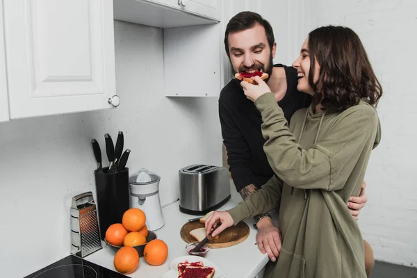 Femme souriante nourrir l'homme avec des toasts pendant le petit déjeuner dans la cuisine — Photo de stock