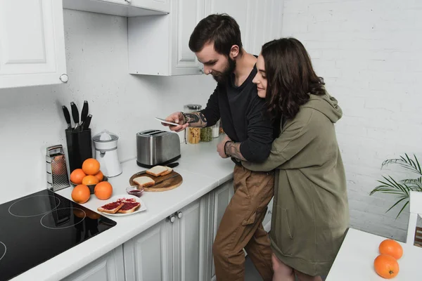 Hermosa pareja joven tomando fotos de brindis en el teléfono inteligente durante el desayuno en la cocina - foto de stock