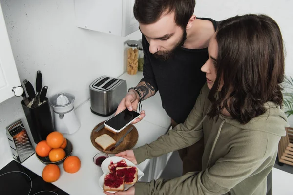 Hermosa pareja joven tomando fotos de brindis en el teléfono inteligente durante el desayuno en la cocina - foto de stock