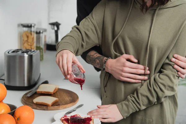 Vista recortada de la mujer que vierte mermelada en tostadas durante el desayuno en la cocina con el hombre en el fondo - foto de stock