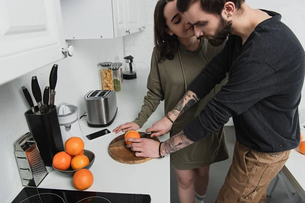 Homem cortando laranja na tábua de madeira com mulher durante o café da manhã na cozinha — Fotografia de Stock