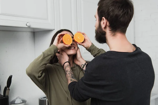 Uomo abbracciando donna che tiene arance davanti al viso in cucina — Foto stock