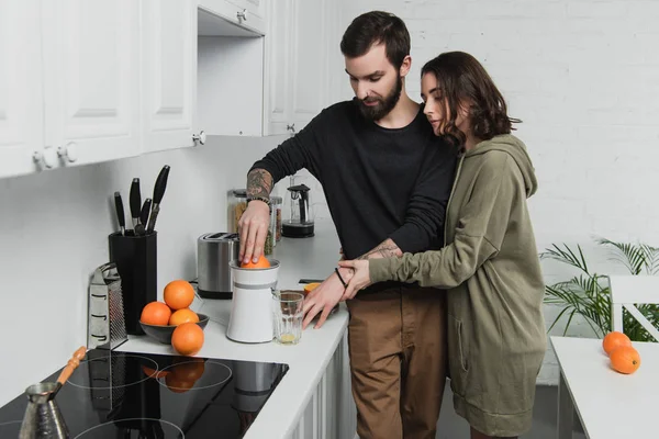 Hermosa pareja joven preparando jugo de naranja durante el desayuno en la cocina - foto de stock