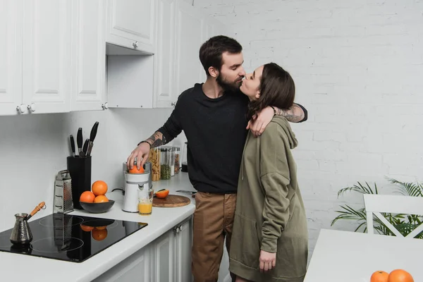 Beautiful young couple making orange juice, hugging and kissing during breakfast in kitchen — Stock Photo
