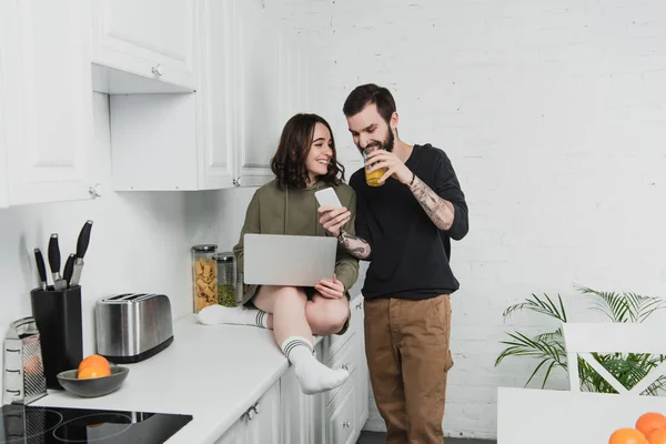 Uomo bere succo di frutta e utilizzando smartphone mentre sorridente donna utilizzando il computer portatile durante la colazione in cucina — Foto stock