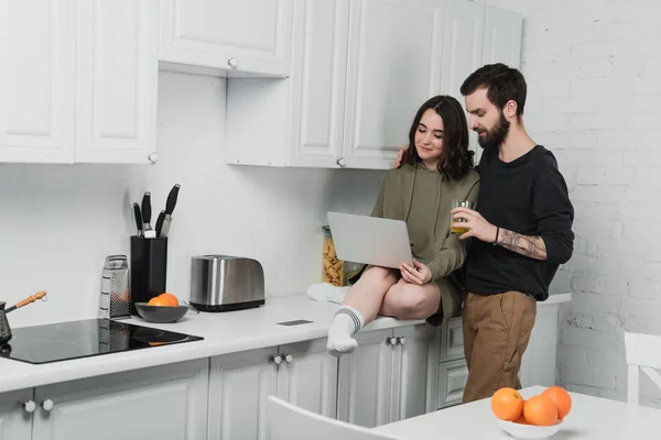 Hombre guapo beber jugo mientras que la mujer usando el ordenador portátil durante el desayuno en la cocina - foto de stock