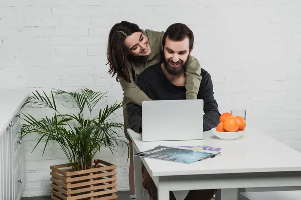 Bella sorridente giovane donna che abbraccia l'uomo felice utilizzando il computer portatile durante la colazione in cucina — Foto stock