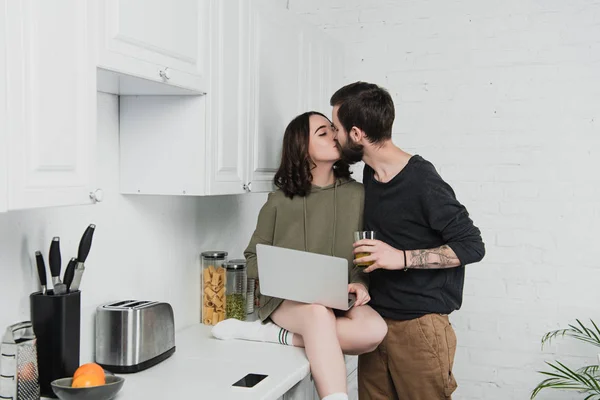 Мужчина целует женщину с ноутбуком во время завтрака на кухне — стоковое фото