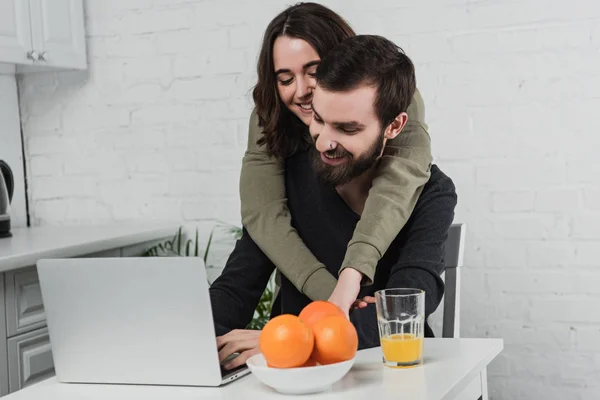 Hermosa mujer joven sonriente abrazando al hombre usando el ordenador portátil durante el desayuno en la cocina - foto de stock