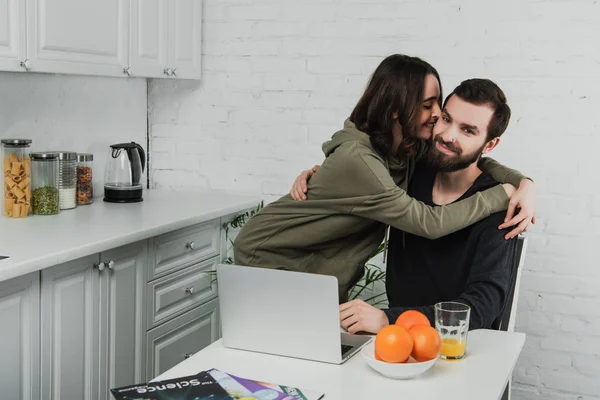 Hermosa mujer joven sonriente abrazar y besar al hombre usando el ordenador portátil durante el desayuno en la cocina - foto de stock
