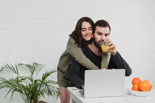 Hermosa mujer joven sonriente abrazando al hombre usando el ordenador portátil y beber jugo de naranja durante el desayuno en la cocina - foto de stock